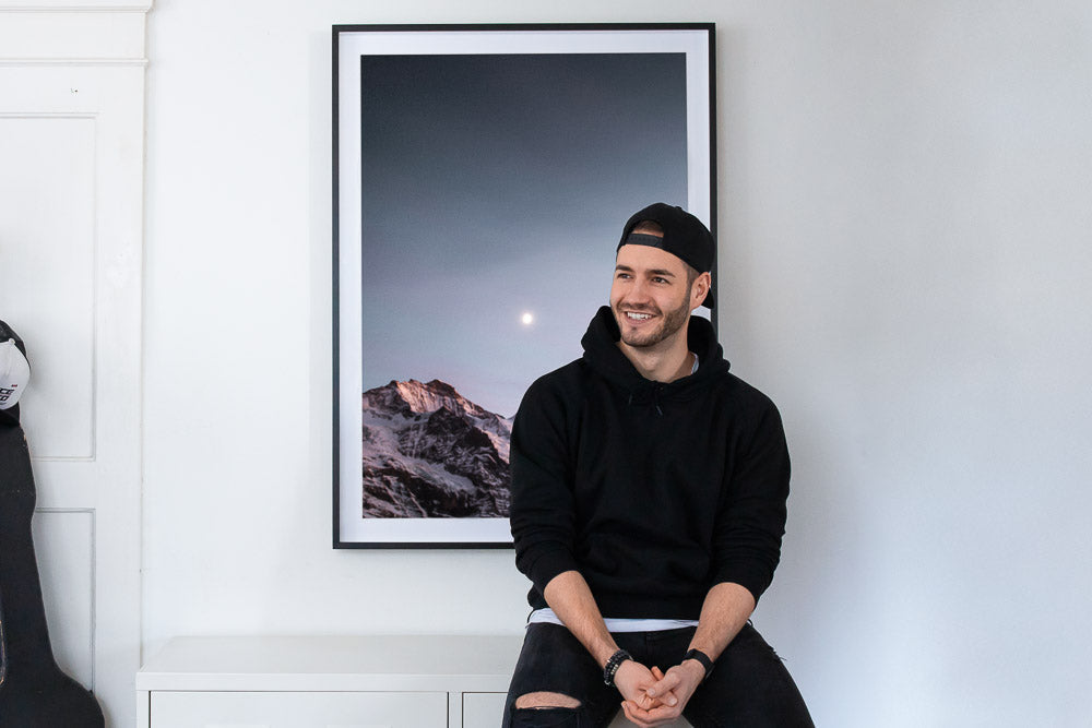 Sandro Breu is smiling in casual black outfit sitting in front of his framed alpine landscape print featuring a snow-capped mountain and rising moon.