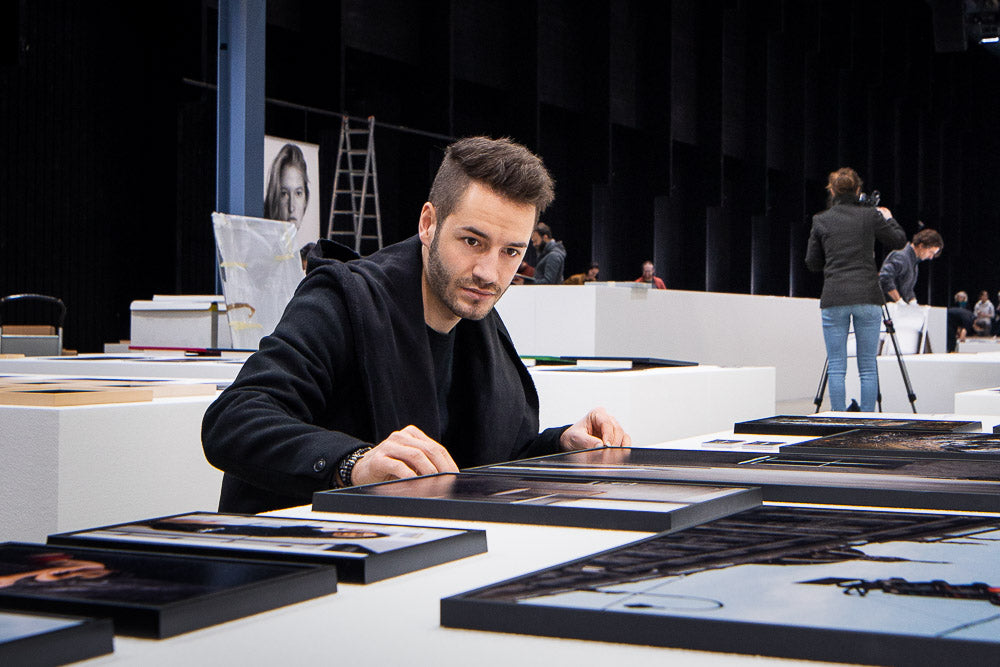 Photographer Sandro Breu reviewing framed prints at an exhibition setup, dressed in a black coat, with a focused expression – behind-the-scenes look at professional curation and artistic presentation.