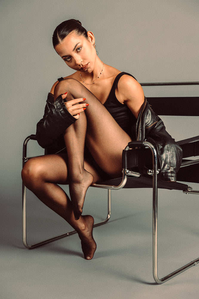 Elegant woman in a black bodysuit and sheer tights sitting on a modern designer chair, draped in a leather jacket. With an intense gaze and poised posture, the portrait exudes confidence and sophistication in a minimalist studio setting.