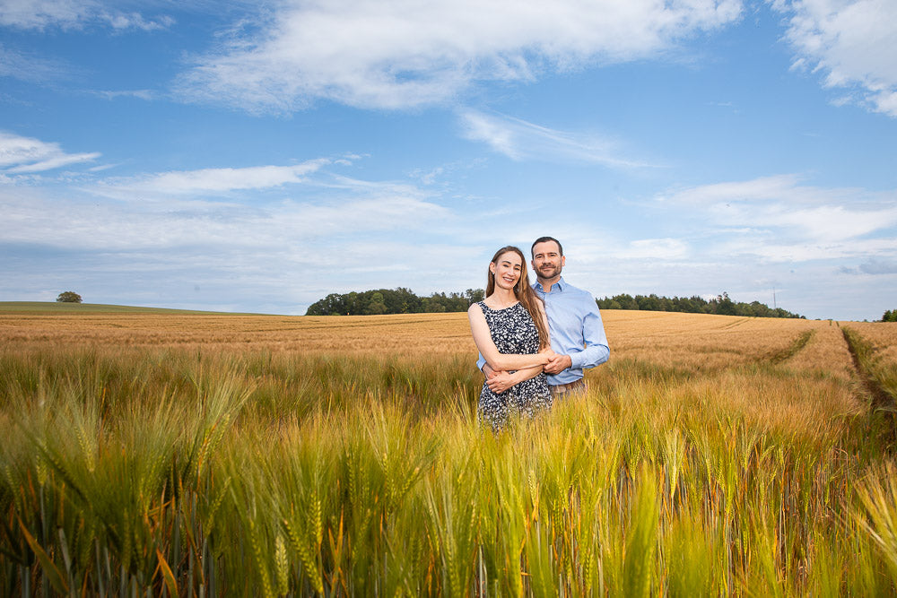 Smiling couple embracing in a golden wheat field under a blue sky, radiating happiness and love. The serene rural landscape and natural sunlight create a warm, romantic atmosphere.