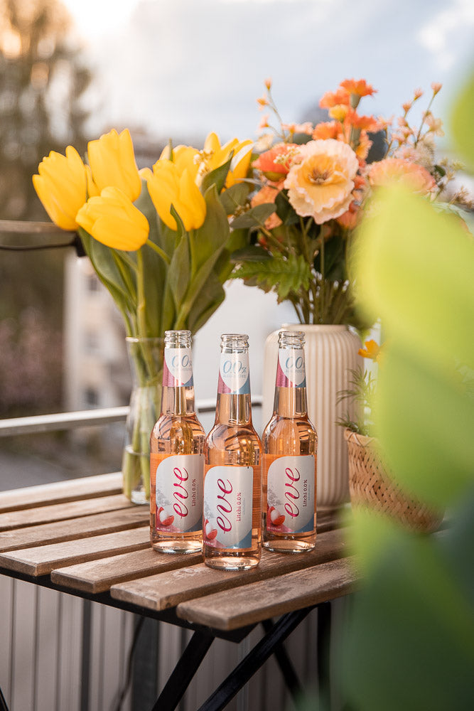 Three Eve Lychee Cooler bottles on a wooden table, surrounded by vibrant yellow tulips and pastel flowers, creating a fresh and inviting springtime atmosphere on a balcony.