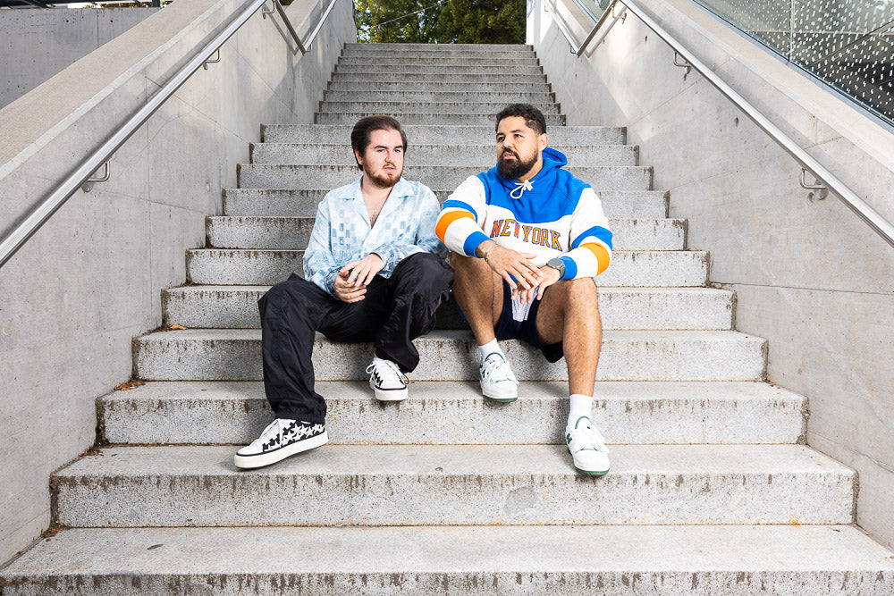 Gil Glaze and Rapture Boy sitting on a concrete staircase in an urban setting, engaged in conversation. One wears a light blue patterned shirt with black pants, while the other sports a bold blue and white 'New York' hoodie with shorts, exuding a modern streetwear vibe.
