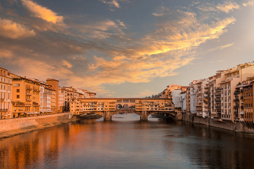 Sunset over the Ponte Vecchio in Florence, Italy, with golden light reflecting on the Arno River. The historic bridge, lined with shops, stands against a dramatic sky, surrounded by picturesque Italian architecture.