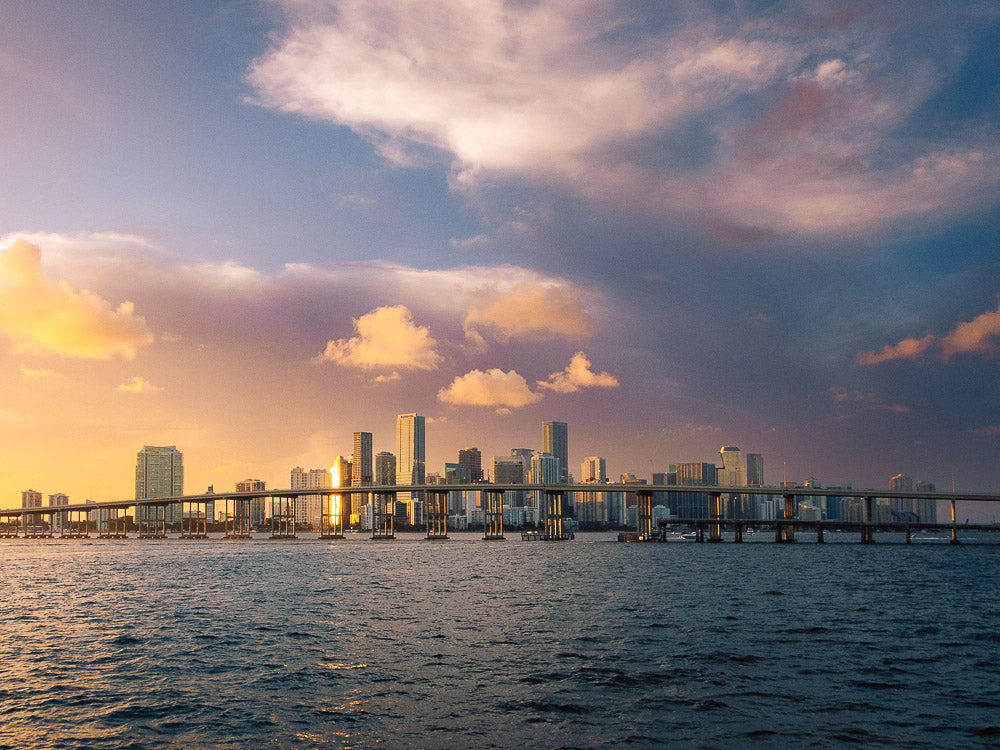 Stunning Miami skyline at sunset with golden light reflecting off the skyscrapers, a scenic bridge stretching over the blue waters, and a dramatic sky with colorful clouds.