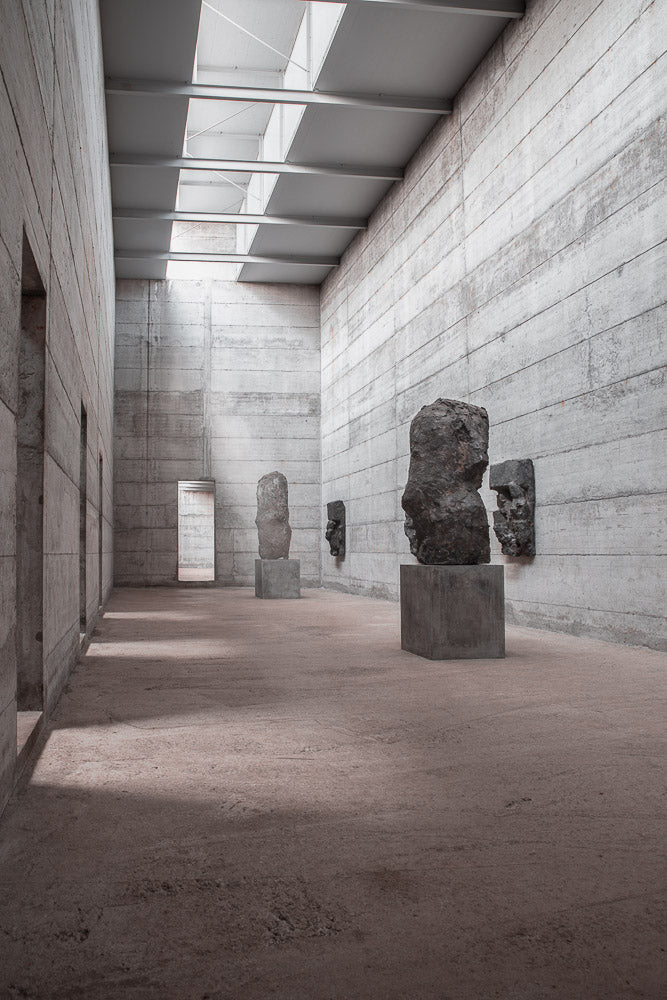 Minimalist concrete museum interior with natural light streaming through skylights, highlighting abstract stone sculptures on pedestals in a serene exhibition space.