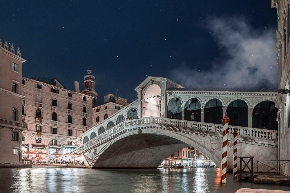 Night view of the iconic Rialto Bridge in Venice, Italy, beautifully illuminated over the Grand Canal, with historic buildings, glowing lights, and a starry sky in the background.