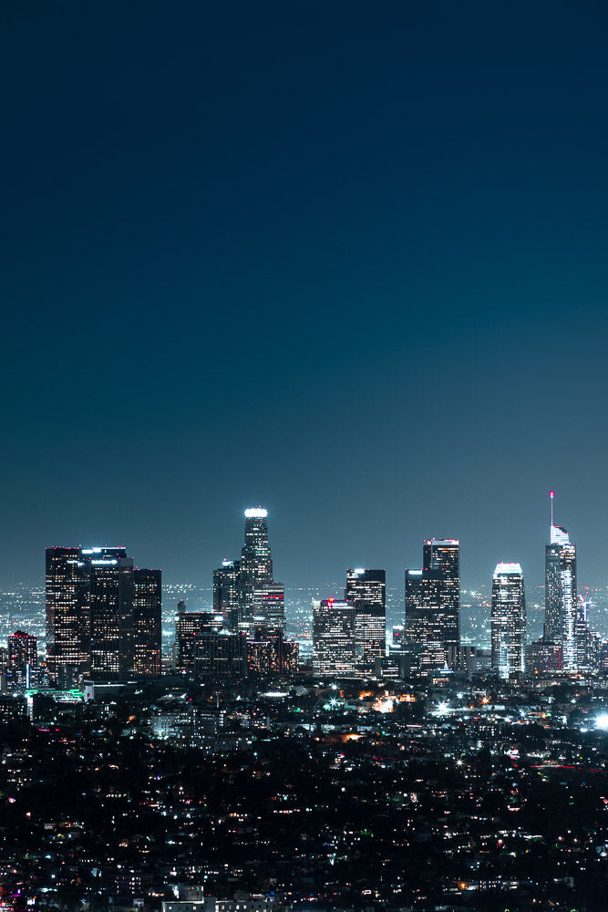 Nighttime cityscape of downtown Los Angeles, California, featuring illuminated skyscrapers and a vibrant skyline against a deep blue sky, captured from an elevated viewpoint.