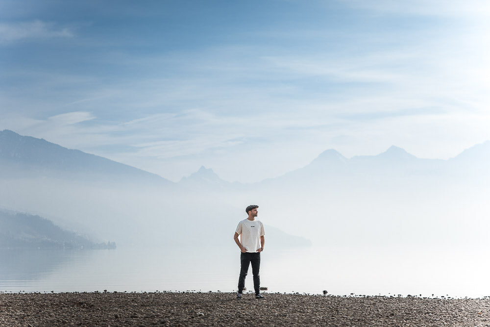 the singer Eifach Ben standing alone on a rocky lakeshore, surrounded by misty mountains and a serene sky, creating a tranquil and contemplative atmosphere in nature.