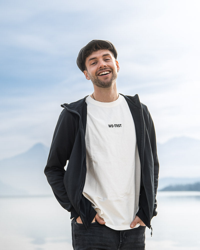 Smiling Eifach Ben in a casual outfit with a 'WE•TRST' t-shirt and a flat cap, standing against a misty mountain and lake backdrop, exuding confidence and positivity.