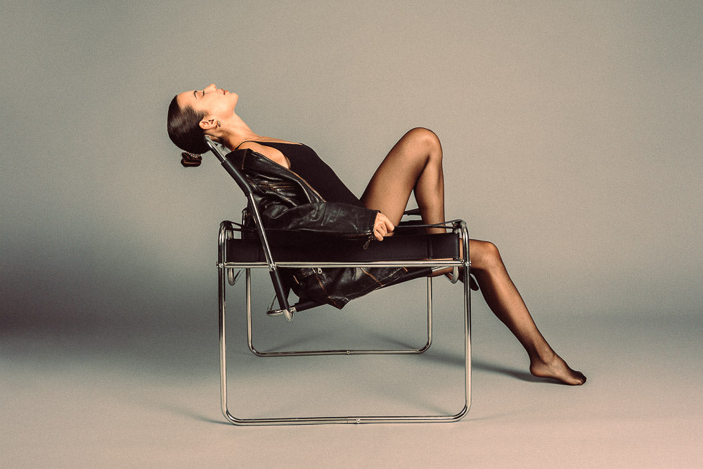 Elegant woman in a black outfit and leather jacket reclining in a modern designer chair, set against a minimalist studio backdrop with a stylish vintage aesthetic.
