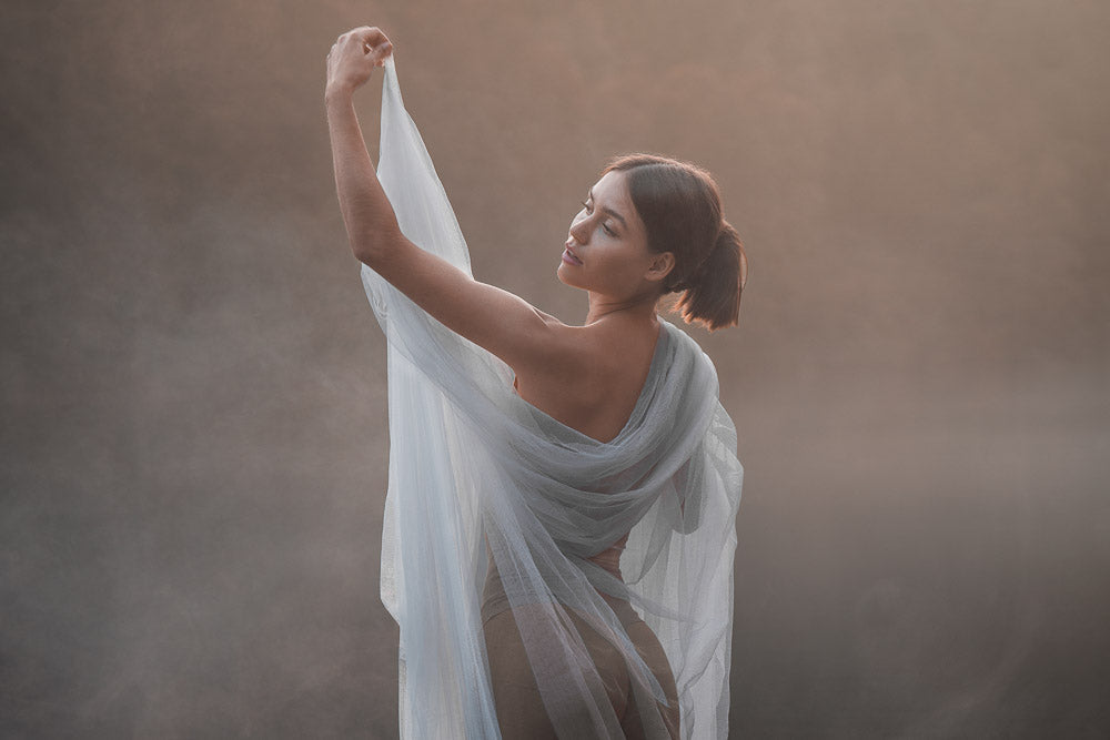 Elegant woman in a sheer, flowing blue fabric poses gracefully in soft natural light, surrounded by mist. Her serene expression and delicate movement create a dreamy, ethereal atmosphere, blending beauty and nature in a fine art portrait.