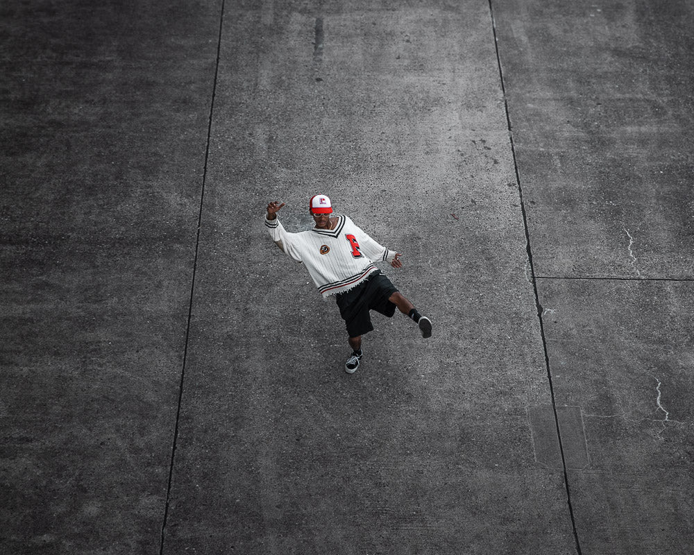 Man in a stylish oversized white sweater with red and black accents, black shorts, and a red baseball cap captured mid-motion on a vast concrete surface. Shot from a high angle, the dynamic pose and urban aesthetic emphasize movement and streetwear fashion.