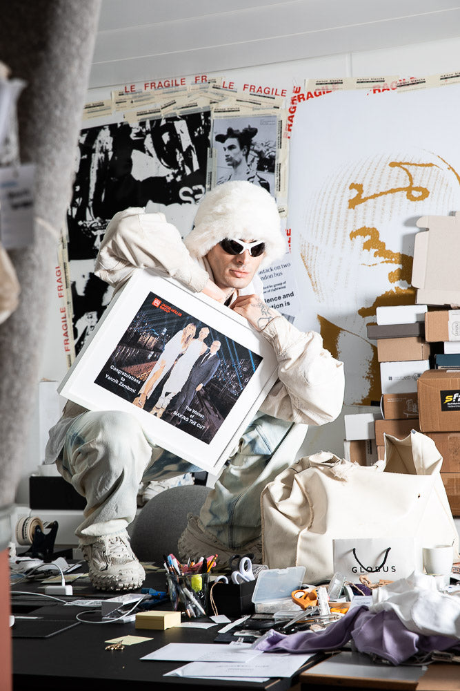 Fashion designer Yannik Zamboni in an eclectic workspace, dressed in an avant-garde white outfit with a fluffy hat and futuristic sunglasses, holding a framed award photo. The background features artistic posters, design materials, and stacked boxes, reflecting a creative and chaotic studio environment.