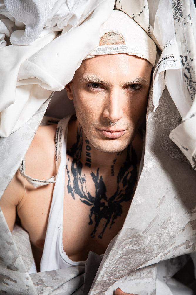 Avant-garde portrait of a male model Yannik Zamboni with bold tattoos, white eyebrows, and a septum piercing, wrapped in textured white fabrics, exuding a high-fashion and artistic aesthetic.