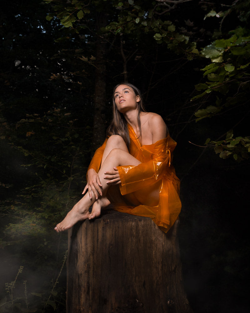 Artistic portrait of a barefoot woman sitting on a tree stump in a mystical forest setting. Wrapped in a translucent orange coat, she gazes upwards, illuminated by soft lighting, creating an ethereal and dreamy atmosphere amidst the dark green foliage.