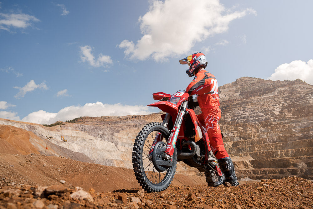 Professional motocross rider Michael Walkner in full red gear sits on a dirt bike in a rugged quarry landscape. The athlete, wearing a Red Bull helmet, surveys the terrain under a bright blue sky, capturing the thrill and adventure of off-road motocross racing.