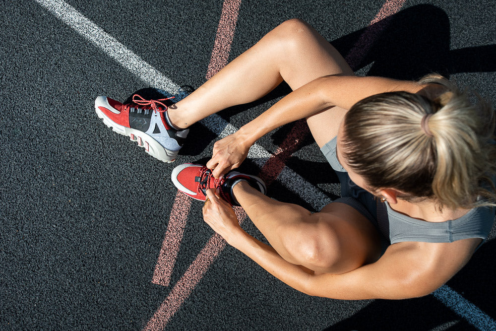 Top-down view of a fit woman tying her red and gray sneakers on a running track, preparing for a workout. A dynamic shot emphasizing movement, athleticism, and sportswear.