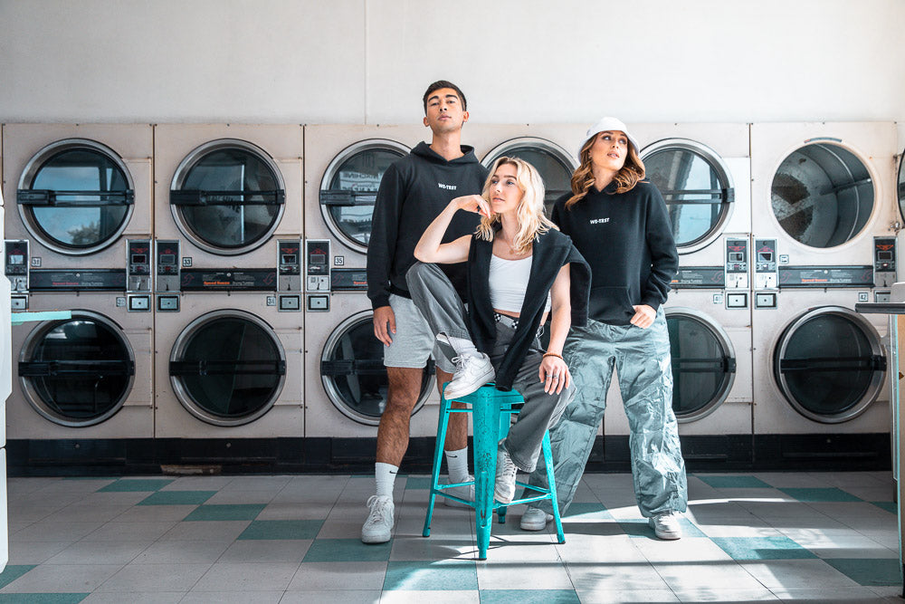 Three models in a laundromat wearing WE•TRST streetwear, featuring black hoodies, cargo pants, and casual shorts – modern urban fashion with a minimalist and bold aesthetic.