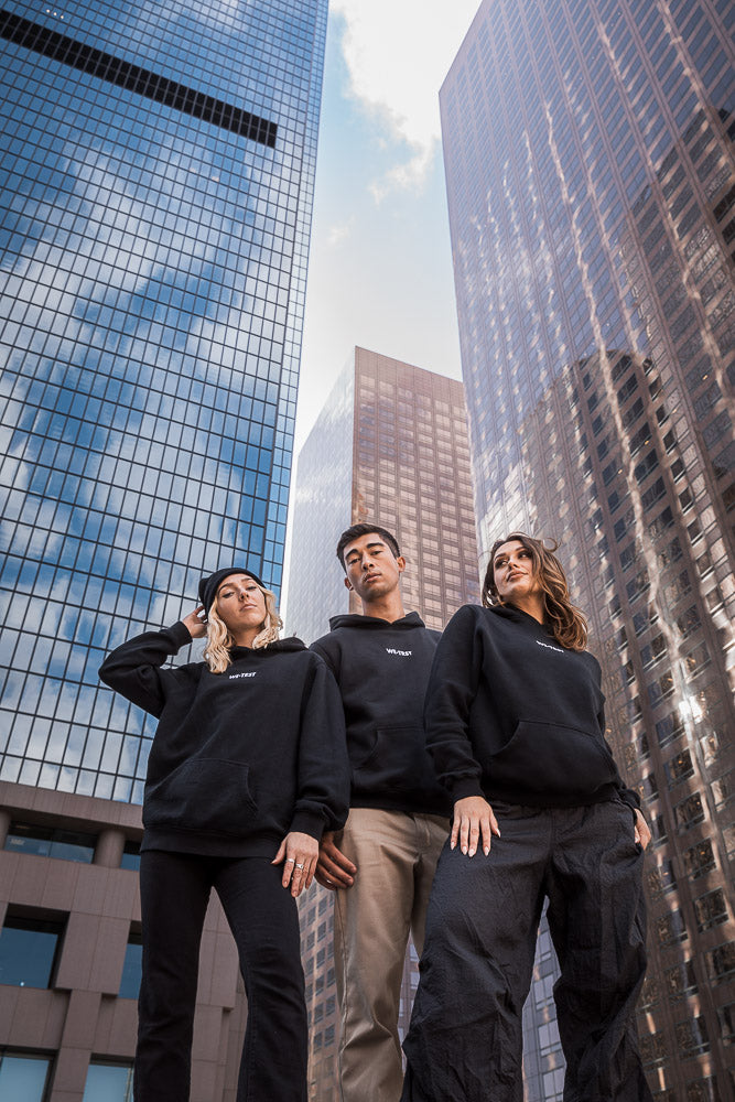 Three models wearing black WE•TRST hoodies pose confidently in an urban cityscape surrounded by towering glass skyscrapers. Streetwear fashion, contemporary style, and urban culture.