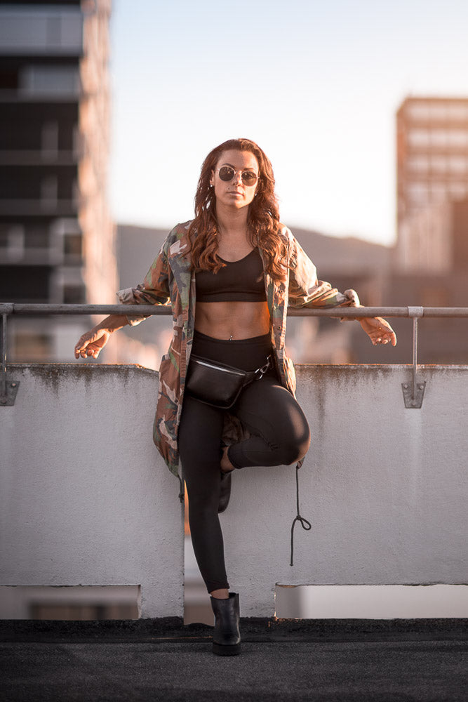 Confident woman in urban streetwear posing on a rooftop at sunset, wearing sunglasses, a camo jacket, black leggings, and a crossbody bag, with a cityscape in the background.