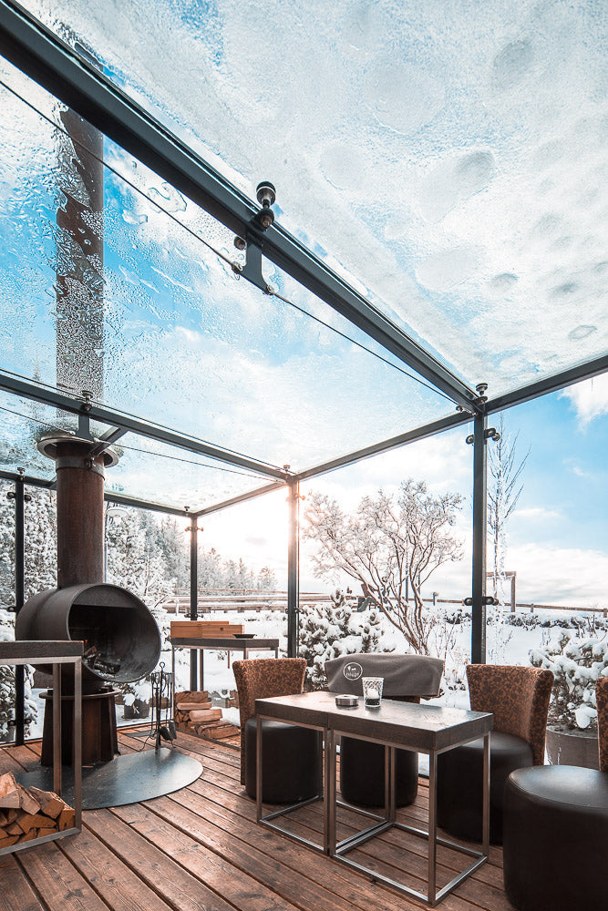 Cozy winter terrace with a glass ceiling covered in snow, featuring a fireplace, modern furniture, and a breathtaking snowy landscape in the background, capturing warmth and luxury in nature