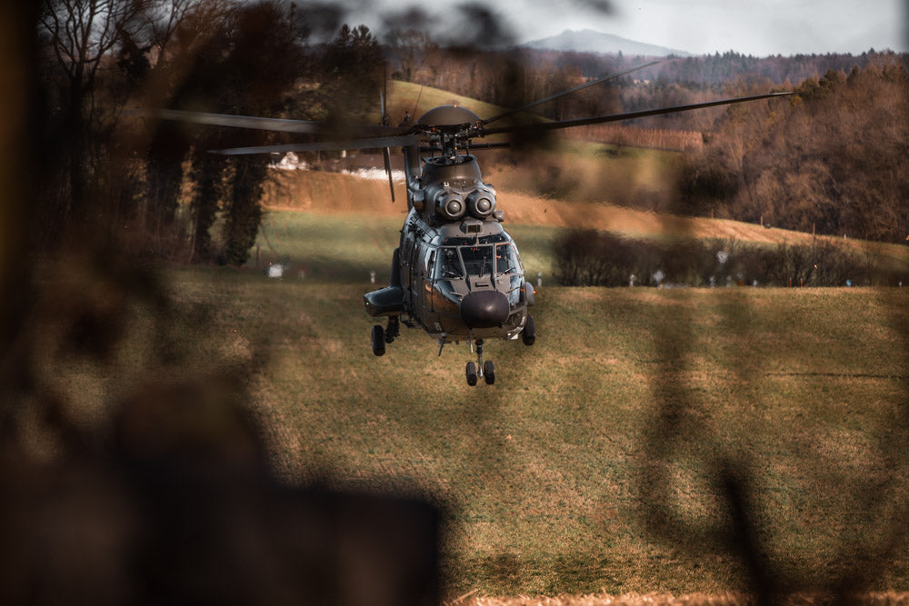 Swiss military helicopter in mid-flight, approaching a rural landing zone with a blurred natural foreground, capturing precision aviation and tactical operations in a scenic landscape.