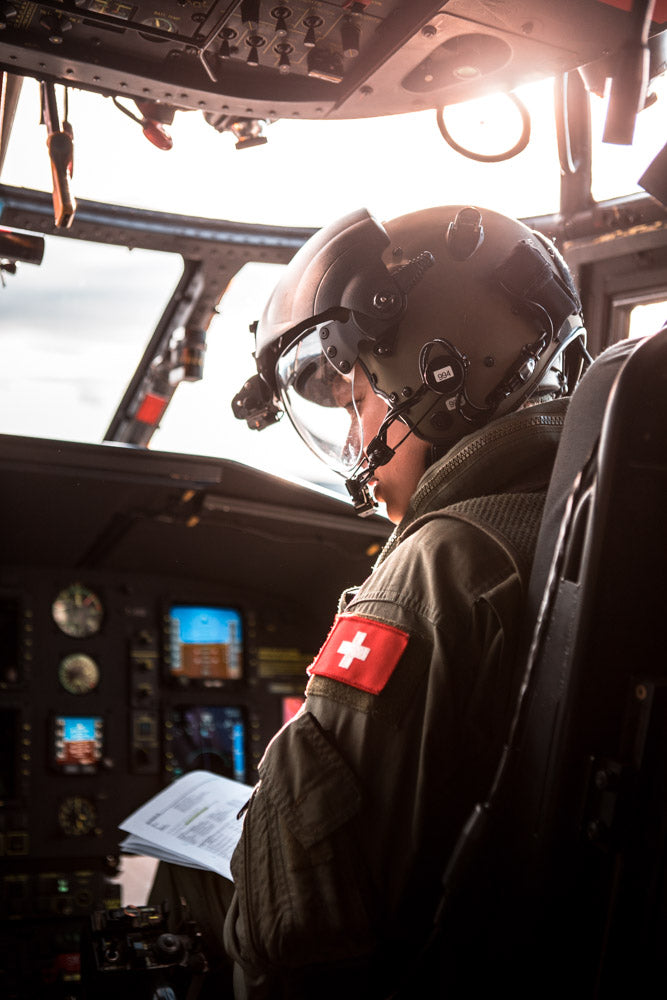 Swiss military pilot inside a helicopter cockpit, wearing a tactical helmet and flight suit with a Swiss flag patch, reviewing flight documents as sunlight streams through the windshield.