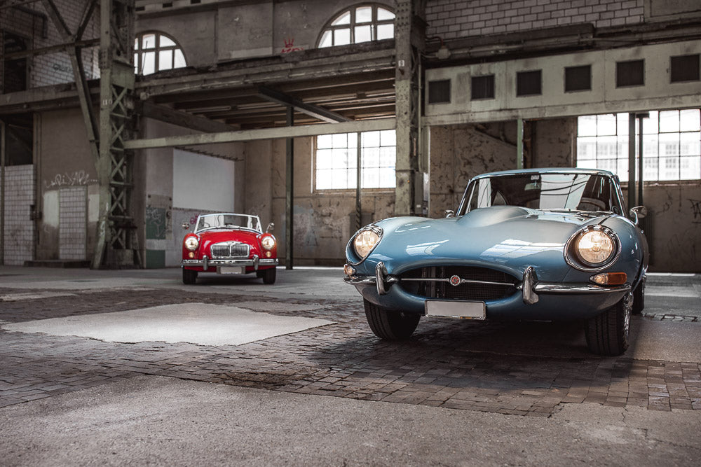 Classic blue Jaguar E-Type and red MG convertible parked inside an industrial warehouse with large windows and metal beams. The vintage cars contrast with the raw, urban backdrop, creating a timeless automotive scene.