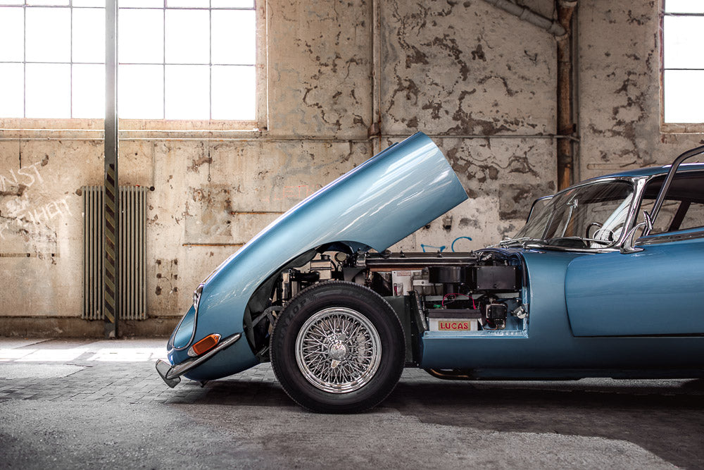 Classic blue Jaguar E-Type with an open hood, showcasing its engine in an industrial warehouse setting. The vintage car’s sleek design and chrome wire wheels contrast with the rustic, weathered walls and large factory windows in the background.