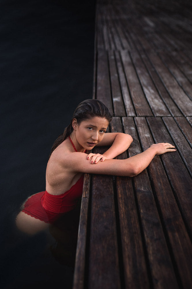 Young woman in a red swimsuit emerging from dark water, leaning on a wooden dock with wet hair and a serene expression, captured in moody natural lighting.