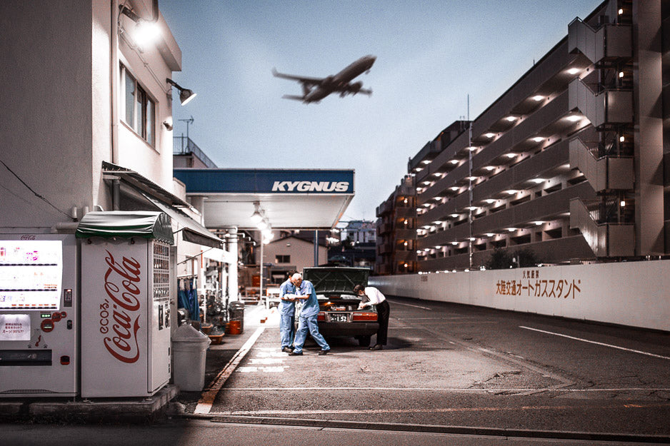 Atmospheric urban scene at a Kygnus gas station in Japan, featuring a vintage car being repaired, a Coca-Cola vending machine, and an airplane flying overhead at dusk.