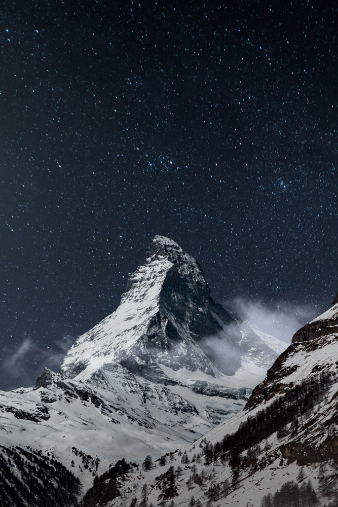 Stunning night view of the Matterhorn mountain in Switzerland, covered in snow and surrounded by a starry sky. The iconic peak stands majestically against the dark alpine landscape, with mist rolling over the slopes.